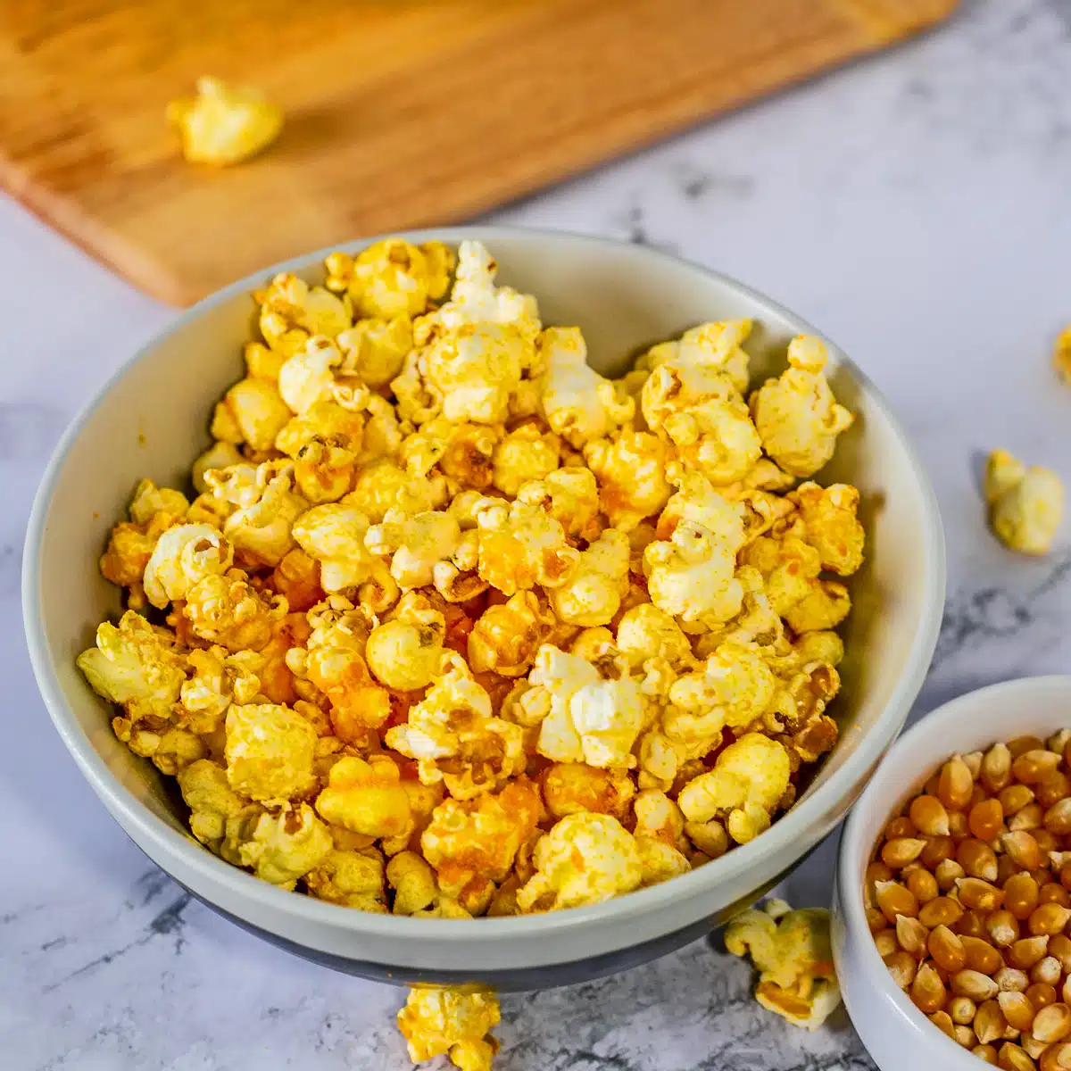 Cheddar popcorn with scattered popcorn on a light marble surface and a bowl of yellow popcorn kernels near it.