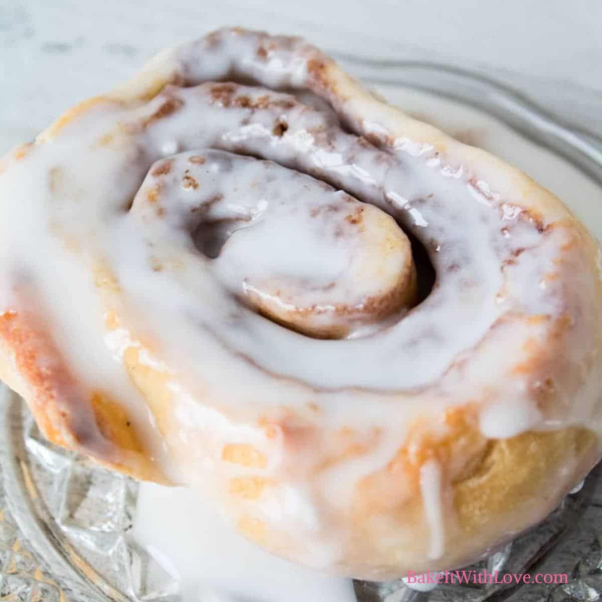 Square image of a cinnamon roll with icing on a glass plate.