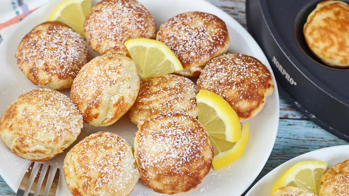 Puffed mini Dutch pancakes or poffertjes plated with lemon and powdered sugar with the pan set to the side.