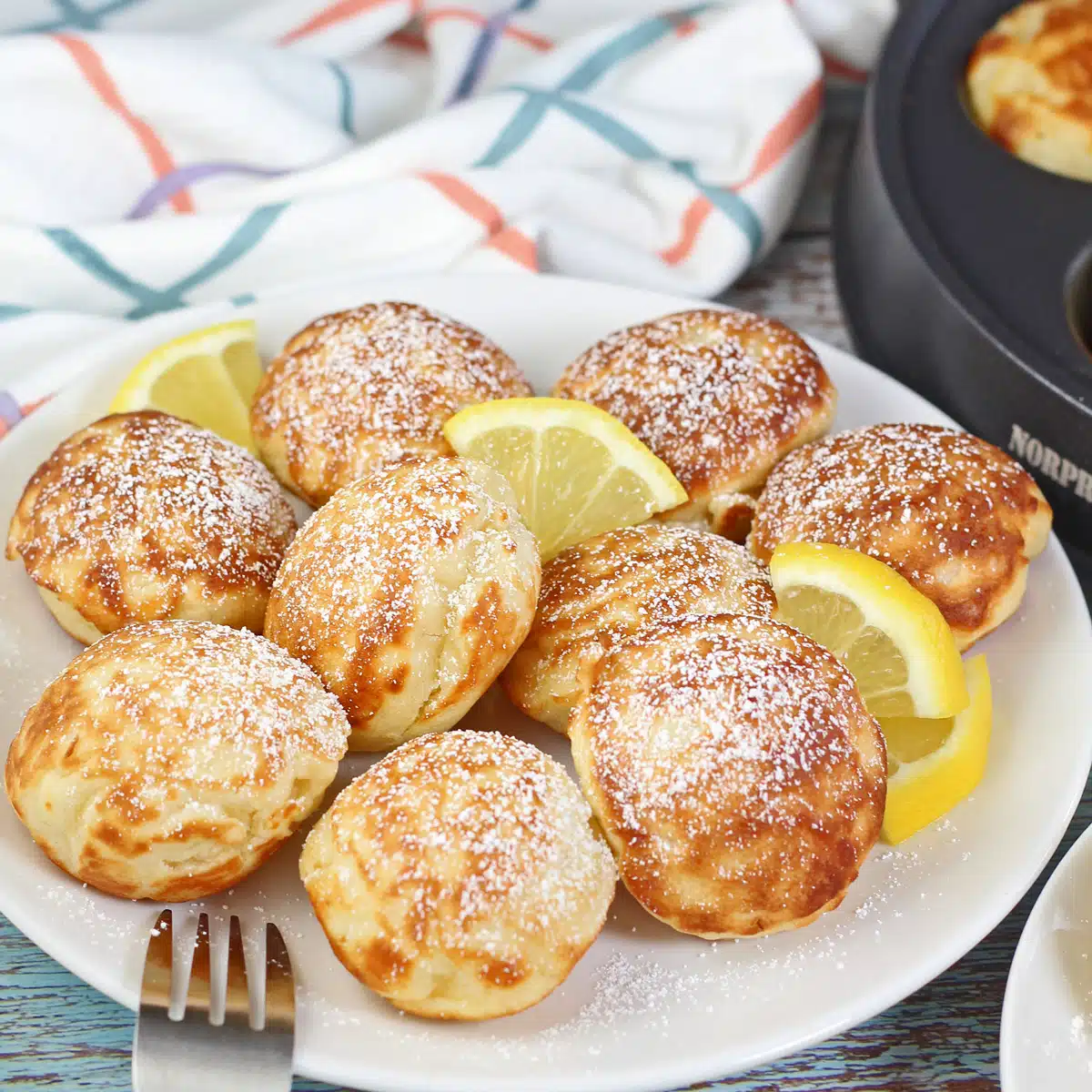 Tasty puffed Dutch pancakes or poffertjes on white plate with small lemon wedges and a dusting of confectioners powdered sugar.