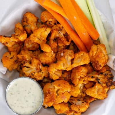 Roasted buffalo cauliflower served in a basket with carrot and celery sticks and some ranch dressing for dipping.