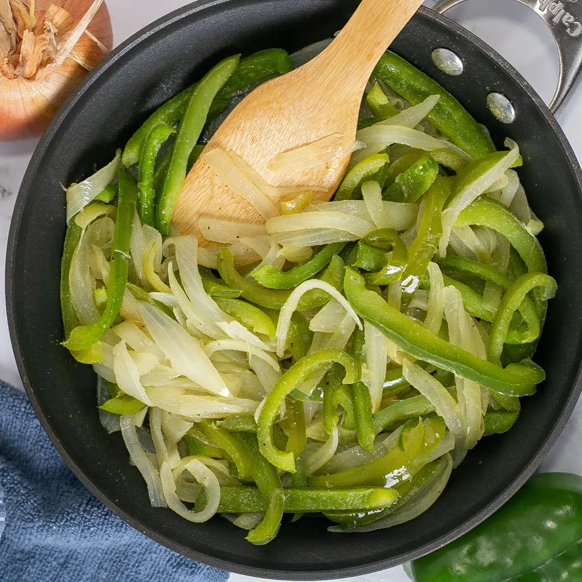 Square image of sauteed peppers and onions in a skillet with a wooden spoon.