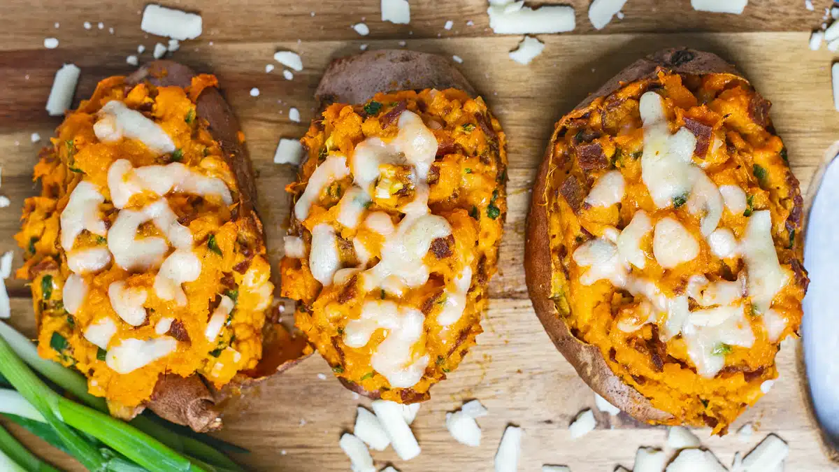 Twice baked sweet potatoes in a row of three on wooden cutting board with green onions on the side and scattered shredded white cheese around them.