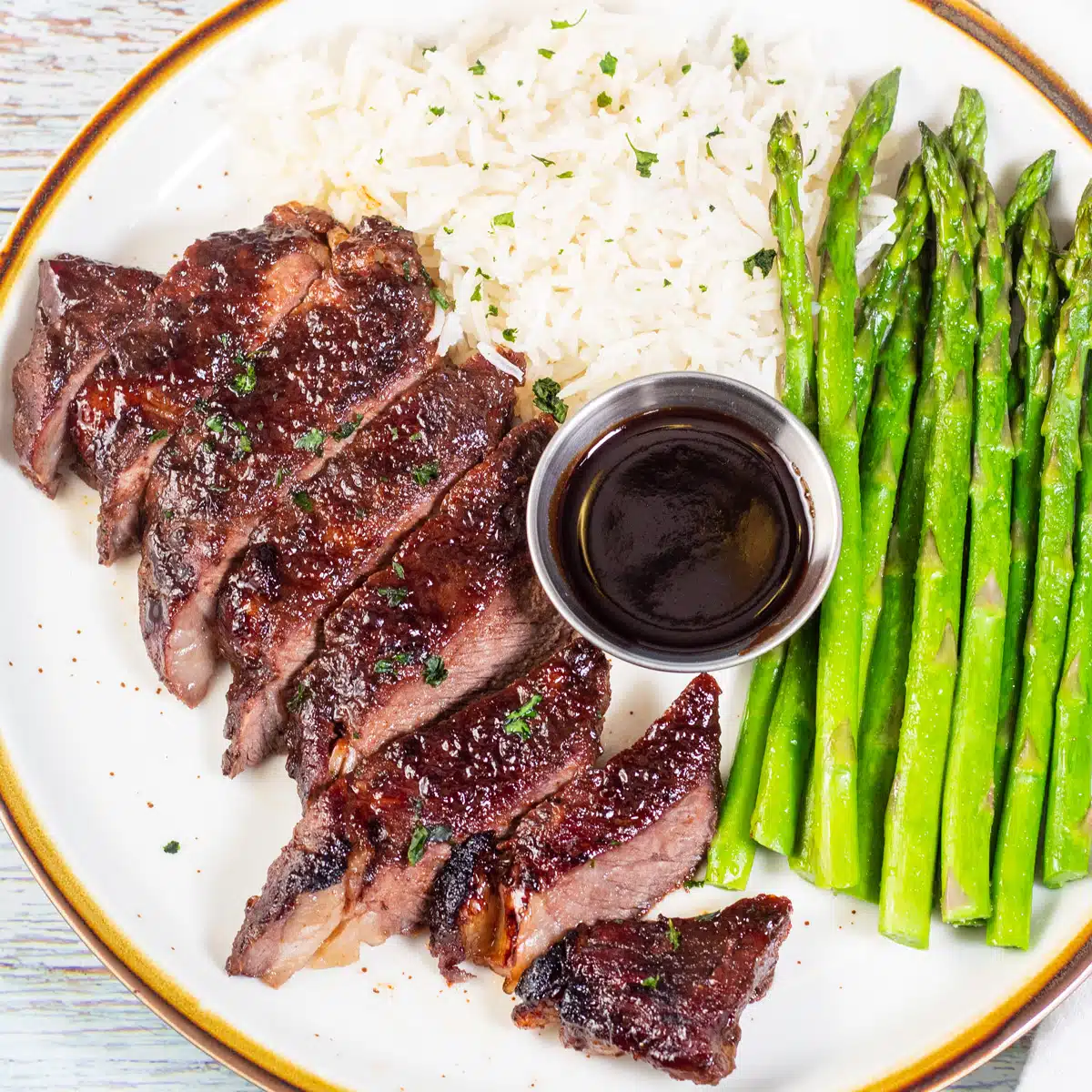 Square image of baked boneless short ribs on a plate with asparagus, rice, and sauce.