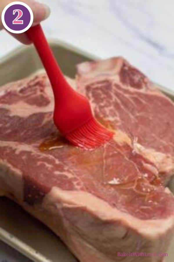 Bistecca alla fiorentina process photo 2. A hand holding a red silicone basting brush is coating a thick cut of raw Bistecca alla Fiorentina (T-bone or porterhouse steak) with oil in a baking pan. A purple circle with the number '2' is in the top left corner.