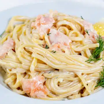 Wide image of creamy smoked salmon pasta on a white plate with dill and a lemon wedge.