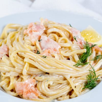 Square image of creamy smoked salmon pasta on a white plate with dill and a lemon wedge.