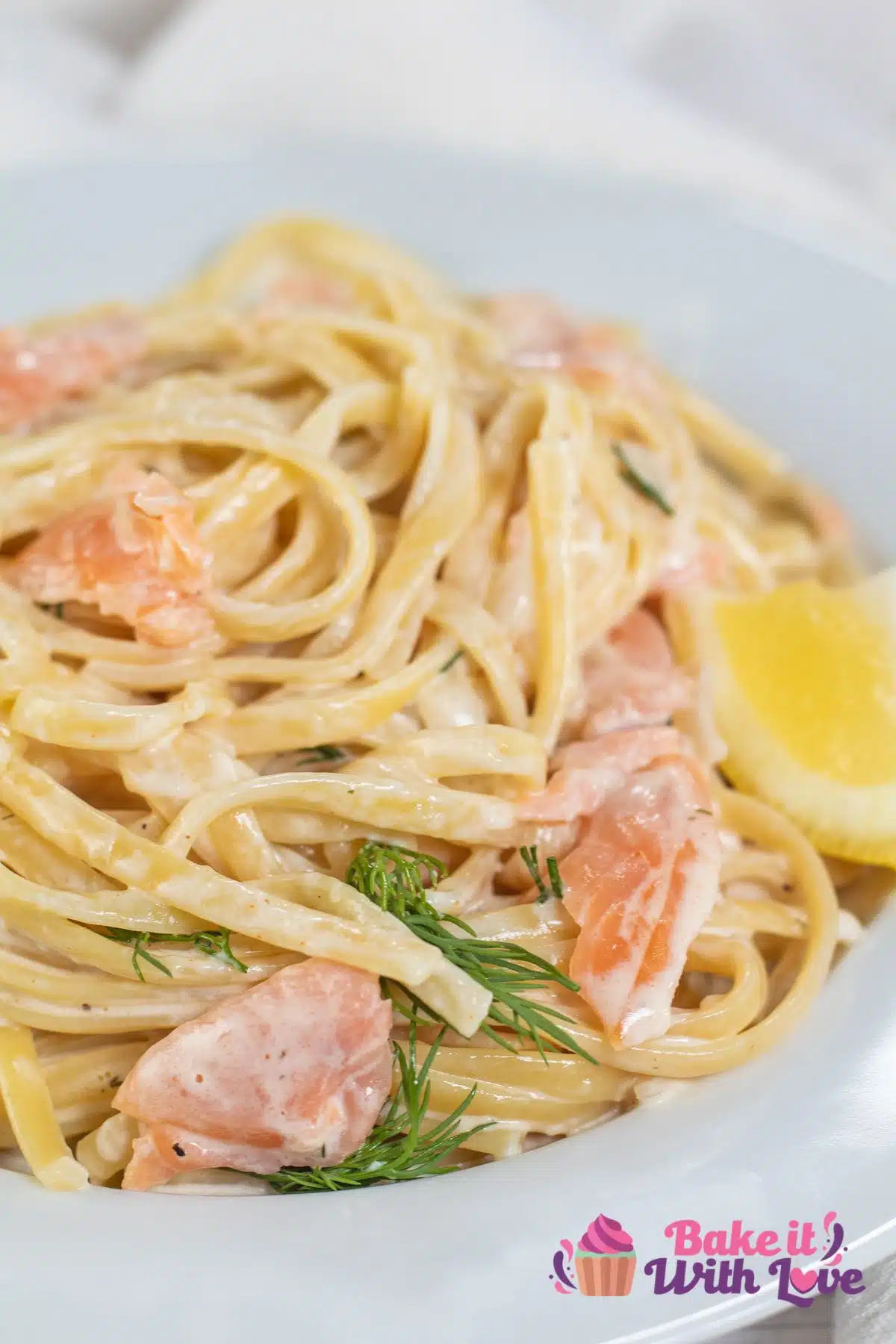 Tall image of creamy smoked salmon pasta on a white plate with dill and a lemon wedge.