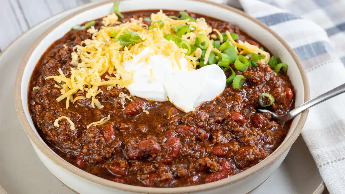 Wide image of Dutch oven chili in a bowl with grated cheddar cheese and sour cream on top.