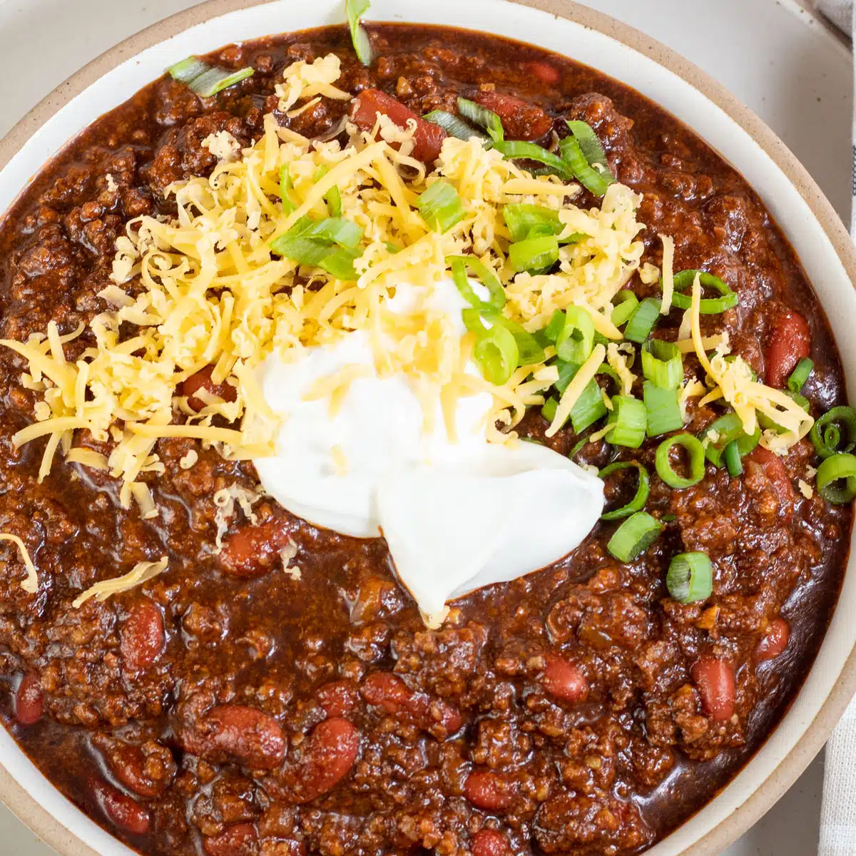 Square image of Dutch oven chili in a bowl with grated cheddar cheese and sour cream on top.