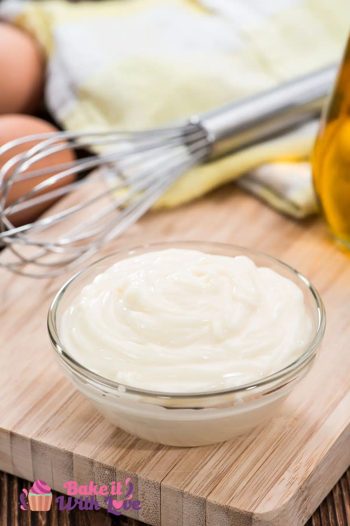 Tasty homemade mayonnaise in a clear glass bowl with a wire whisk and ingredients in the background.