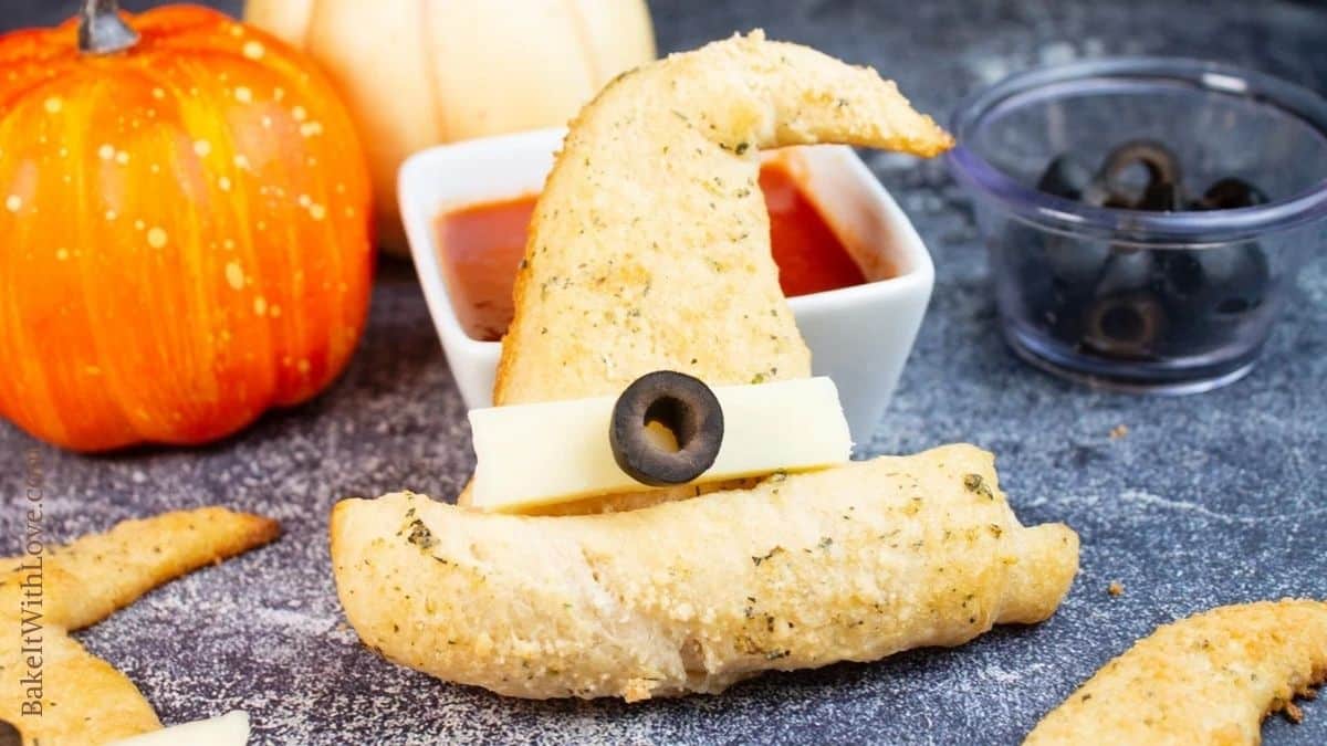 Image of a Halloween crescent roll witches hat leaning against a ramekin of marinara sauce with black olives and a pumpkin in the background. 