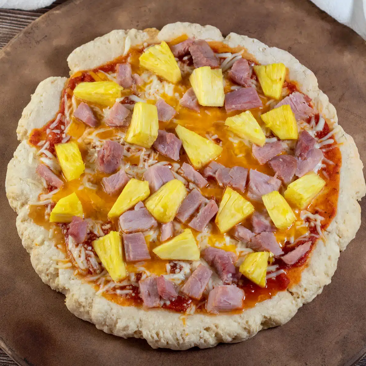 Quadratisches Bild, das eine Pizza im hawaiianischen Stil zeigt, die auf einem Pizzateigboden mit zwei Zutaten zubereitet wird.
