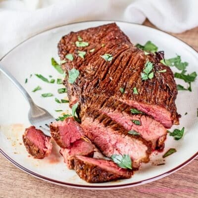 A square image showing tender marinated Denver steak sliced on a plate, served medium rare with a fork and a sprinkle of chopped parsley for a flavorful, steakhouse-style meal.