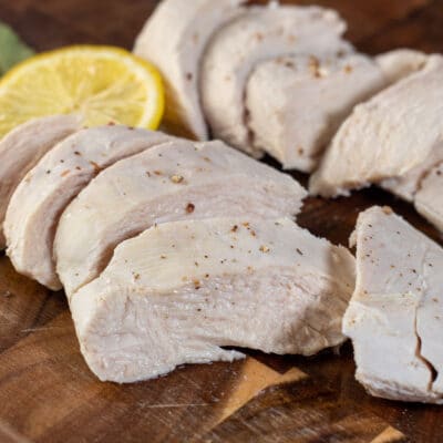 Tender, juicy poached chicken breasts are sliced and shown on a dark wooden cutting board with lemon.