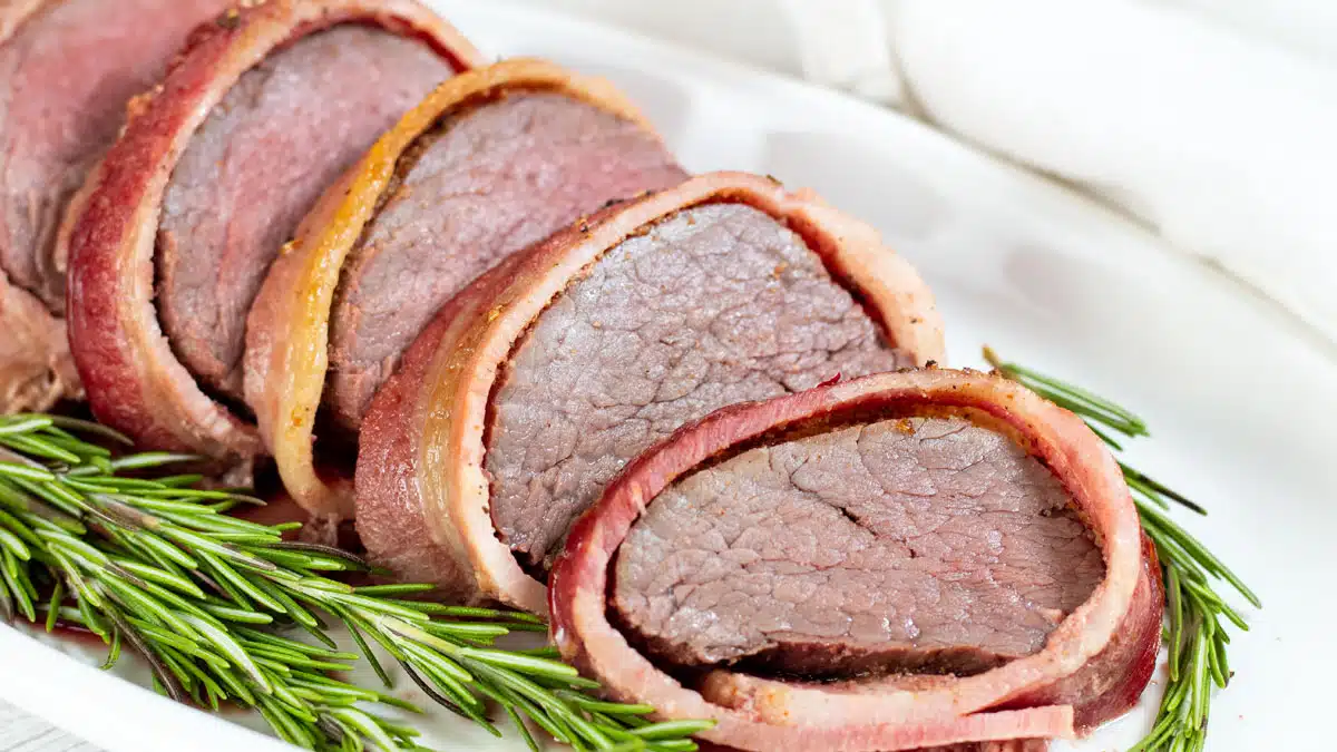 Wide image of bacon wrapped beef tenderloin, sliced.