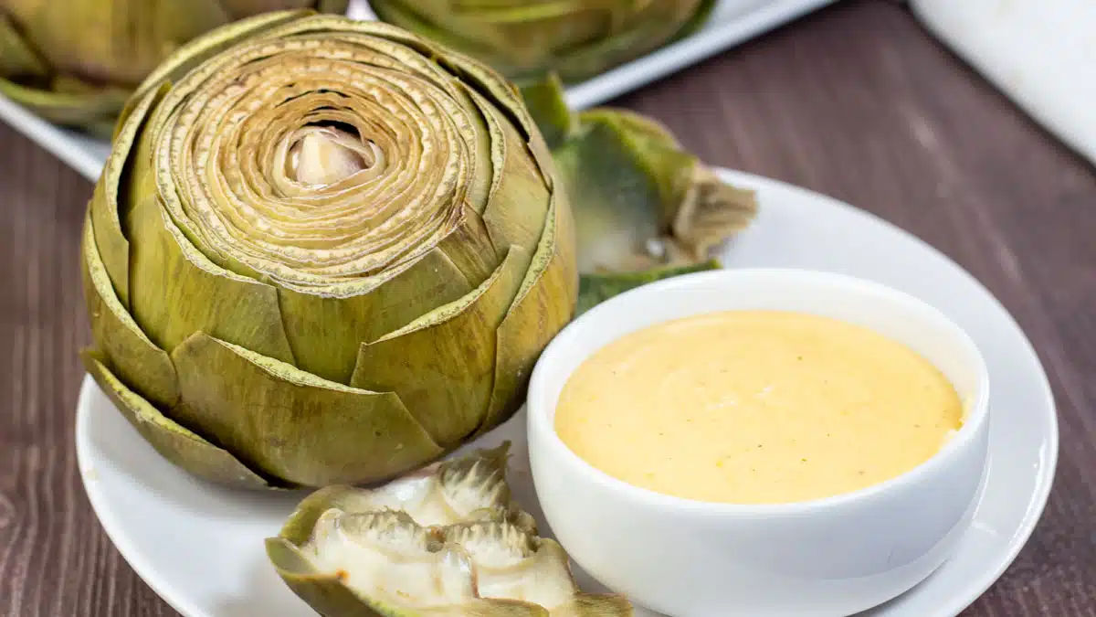 Wide image of steamed artichoke on a white plate with cheese dipping sauce.