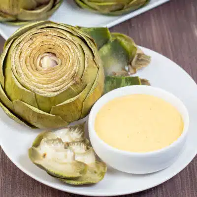 Square image of steamed artichoke on a white plate with cheese dipping sauce.