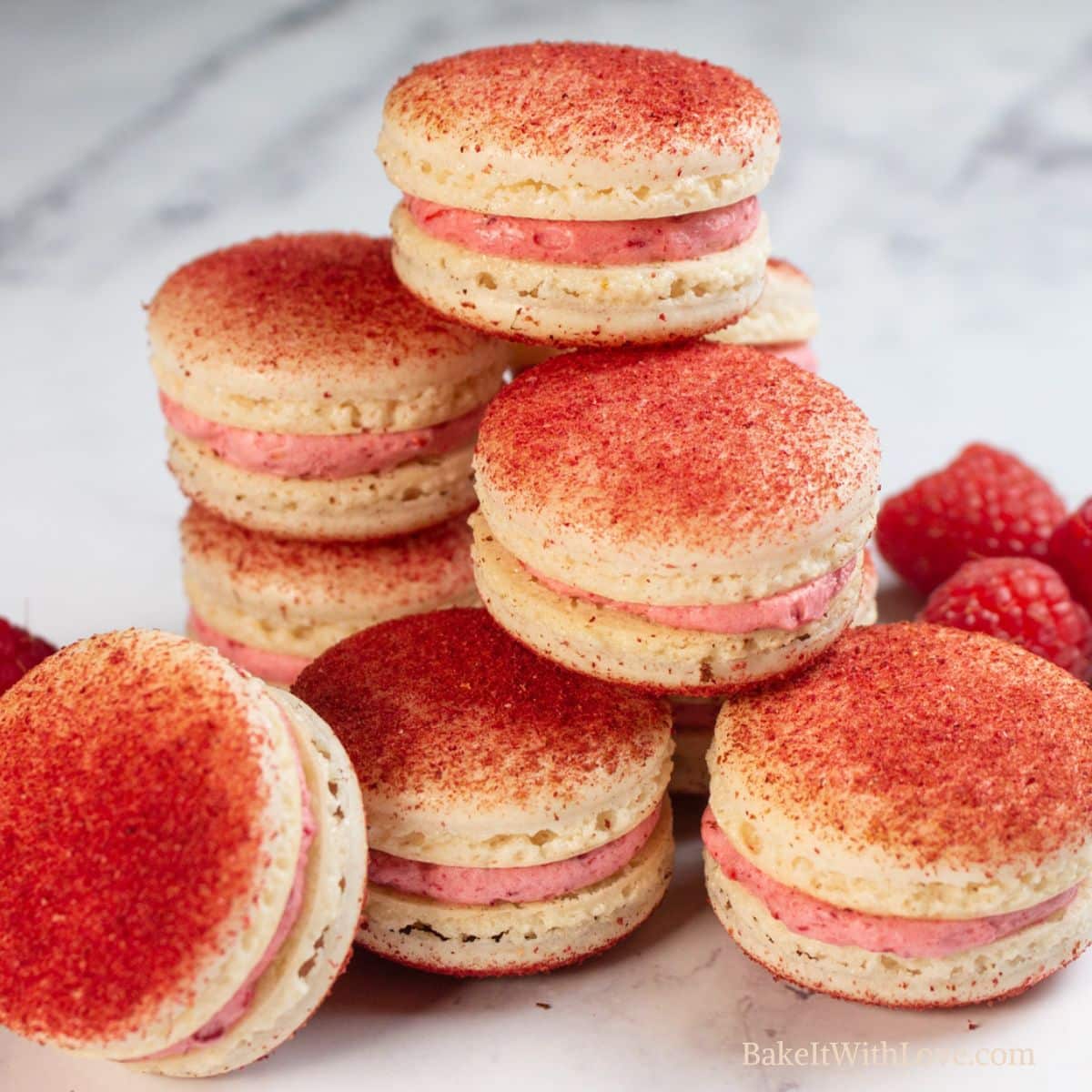 Best raspberry macarons stacked on a marbled grey surface with some fresh raspberries scattered around them.