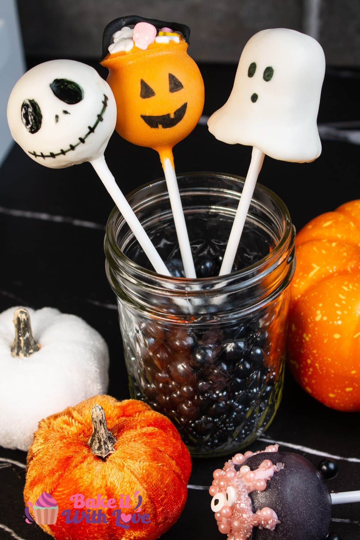 Halloween cake pops propped up in a glass jar filled with black sprinkles and arranged with mini pumpkins around it.