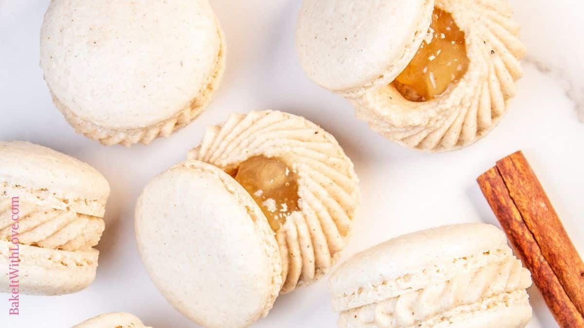 Wide image of apple pie macarons on a marbled surface, some having the tops open so you can see the apple pie filling with a cinnamon stick on the side. 