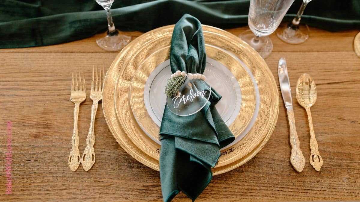 Wide image showing a wooden table with a place setting showing a stack of golden plates, golden silverware, and a green napkin adorned with a handmade name tag. 