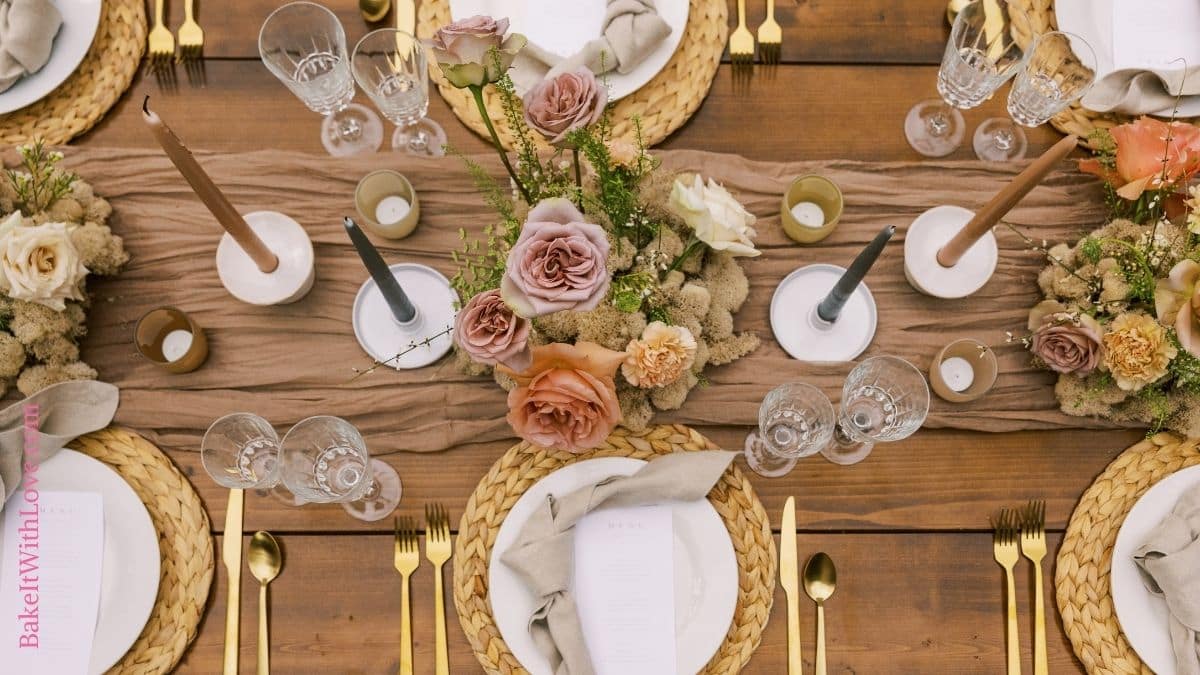 Overhead view of a wooden table with flowers and candles as a centerpiece, charges, plates, napkins, and gold cutlery. 