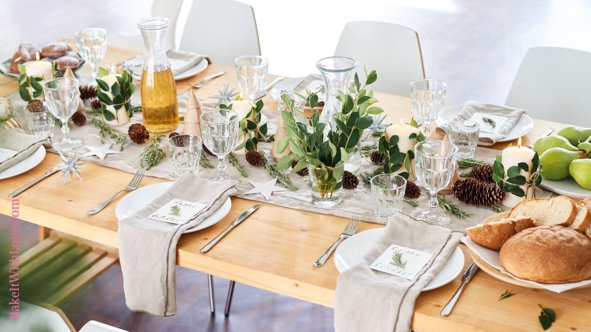 Wide image of a long wooden table with a fabric runner covered in various greenery, glasses, pinecones, candles, and place settings with plates and napkins. 