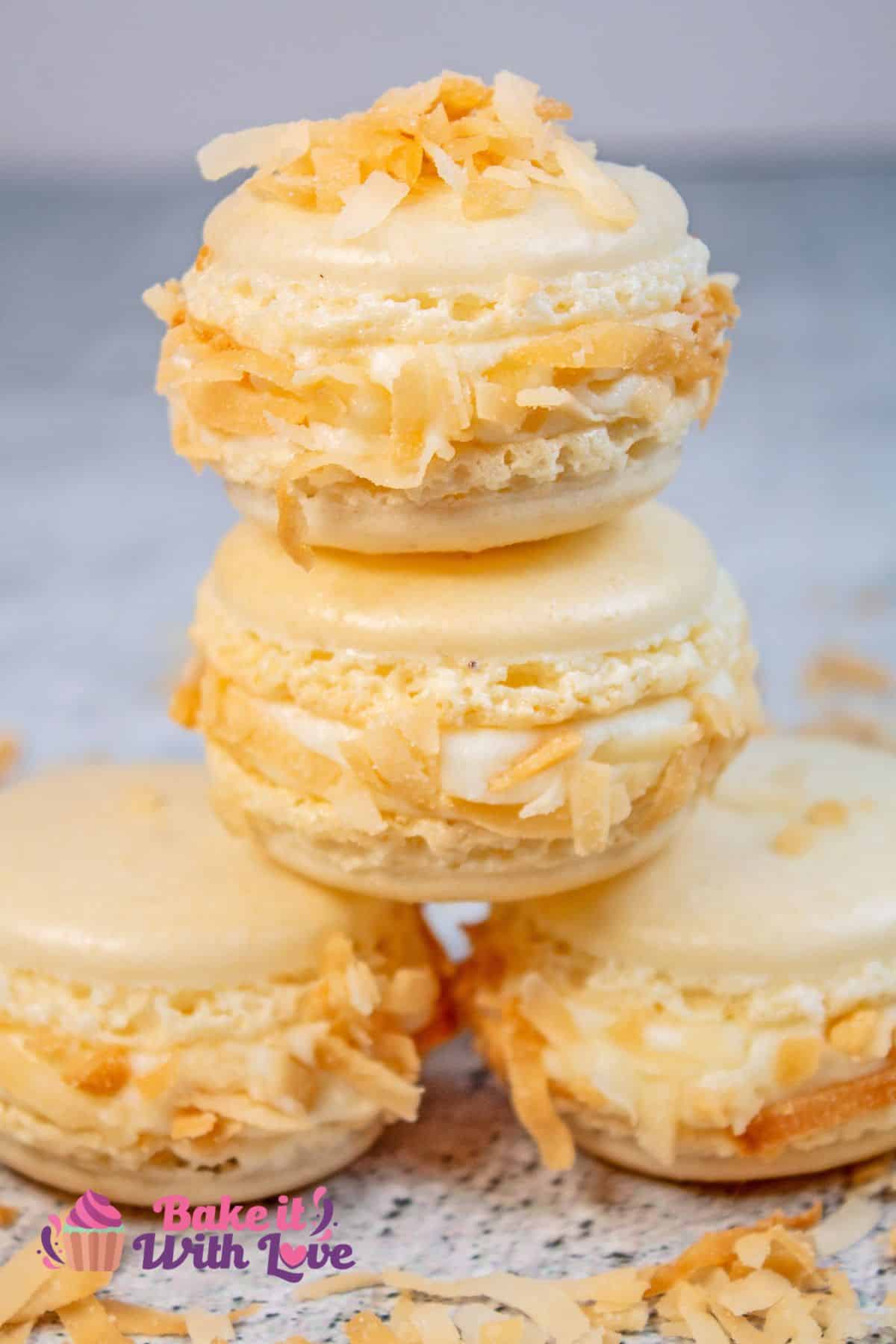 Tall image showing toasted coconut macarons stacked on top of one another and a sprinkle of toasted coconut on top.