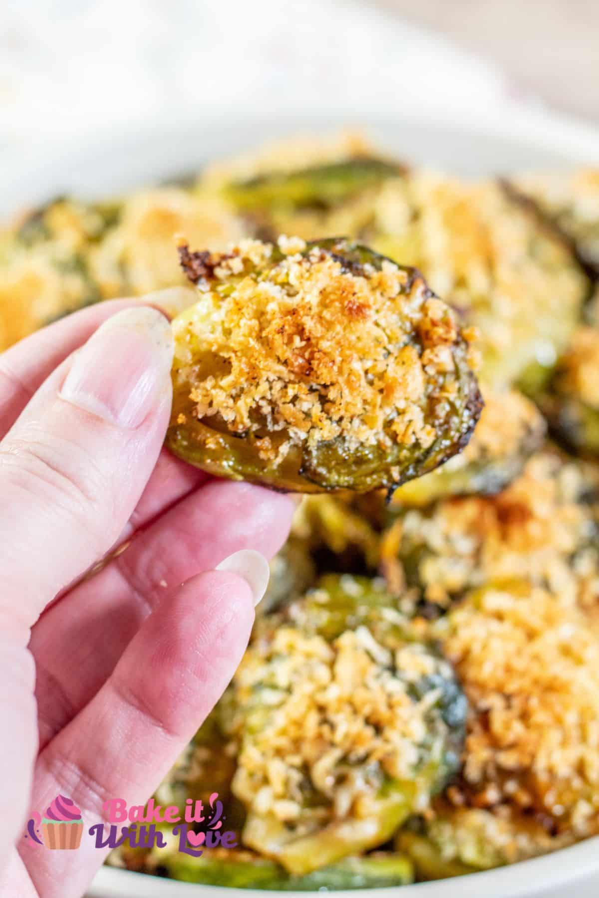 Tall image showing a hand holding up one parmesan smashed brussel sprouts with a bowl full of them in the background. 