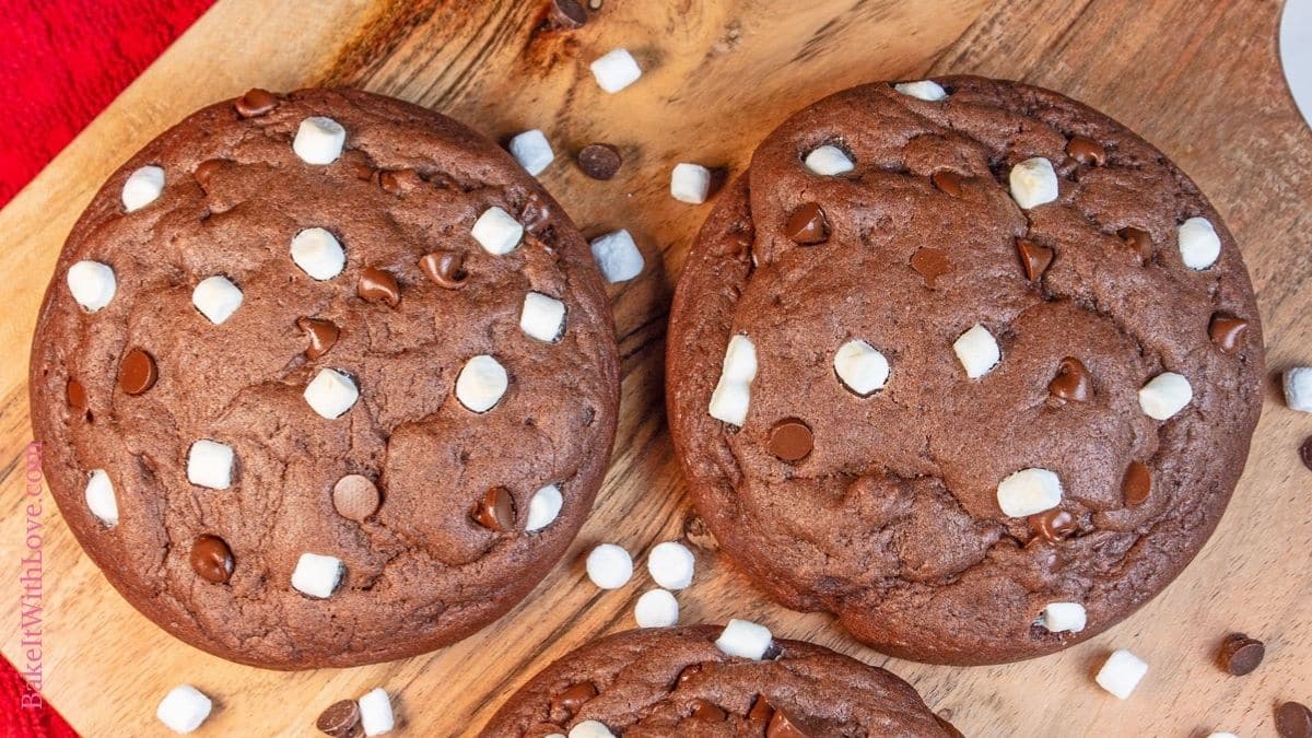 Wide image showing two hot chocolate cookies side by side on a wooden surface surrounded by chocolate chips and mini marshmallow bits. 