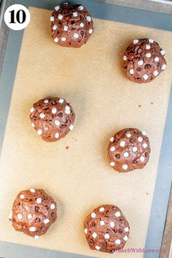 Hot chocolate cookies process photo 10 showing unbaked hot  chocolate cookie dough balls topped with marshmallows and chocolate chips on a silicone baking mat. 