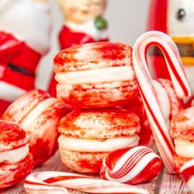 Square image showing a stack of peppermint macarons on a wooden surface with a candy cane leaning against them.