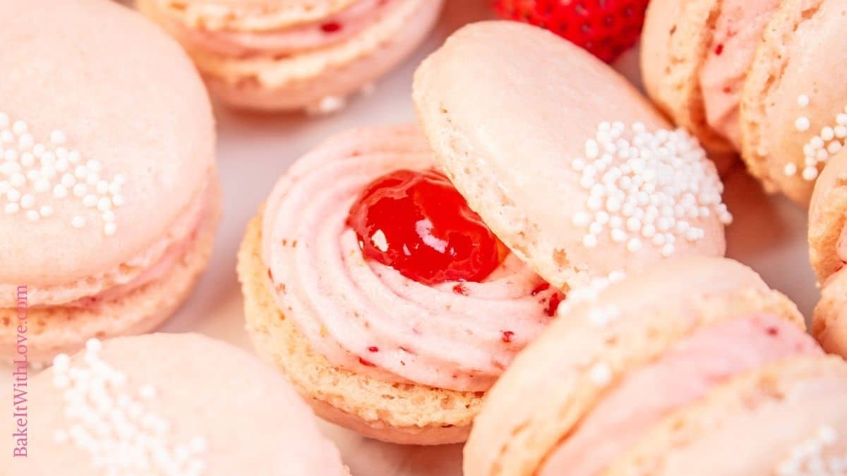 Wide image showing a strawberry macaron with the top shell askew so you can see the frosting and jam filling surrounded by more macarons. 