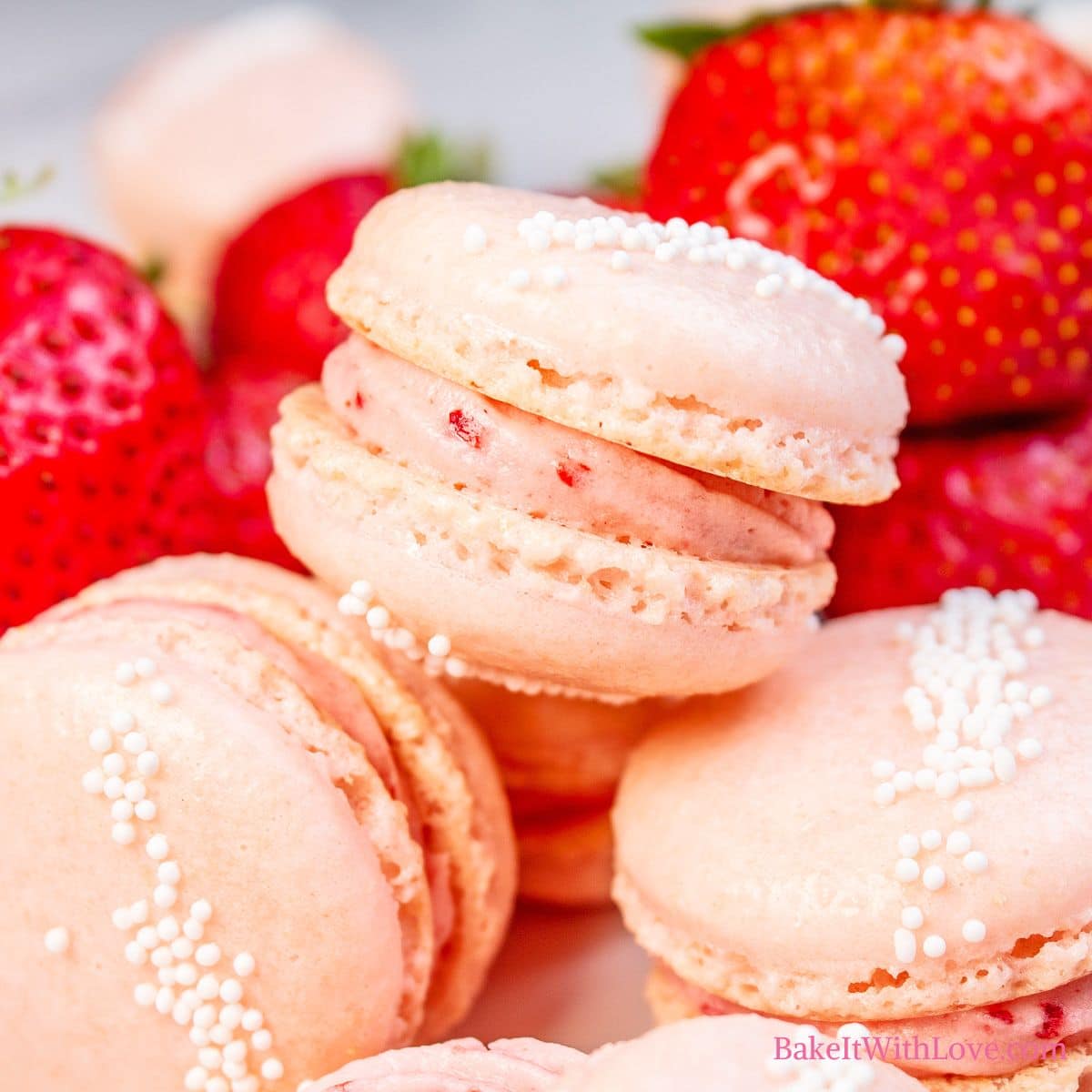 Square image showing a stack of strawberry macarons with fresh strawberries in the background. 