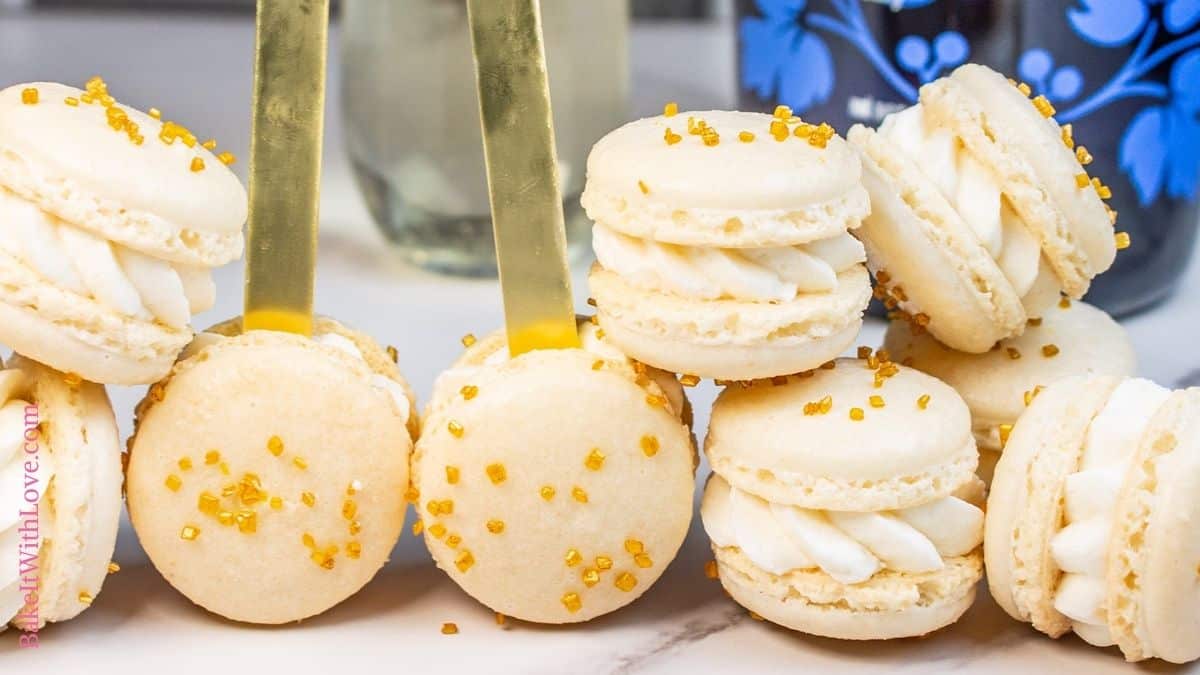 Gold sprinkled champagne macarons stacked randomly on a marbled surface, some with a treat stick inserted to make them 'macaron pops'.