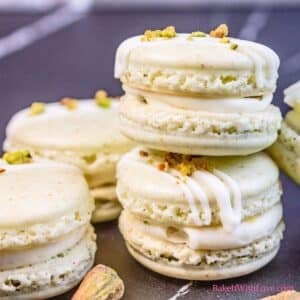 Square closeup on the assembled pistachio macarons on a dark grey background with white chocolate drizzle and sprinkled ground pistachio on the top shells.