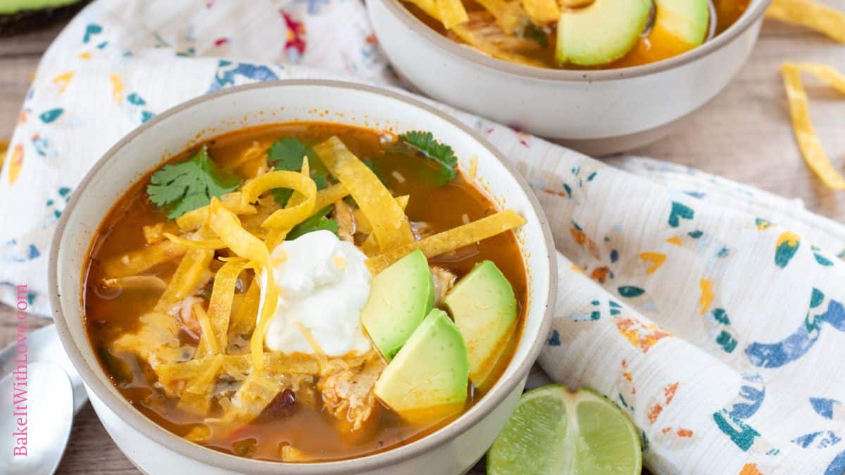Tasty chicken tortilla soup in a tan bowl topped with diced avocado, sour cream, and fried tortilla strips.