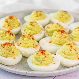 A close-up, slightly elevated shot shows a platter of avocado deviled eggs. Approximately a dozen halved hard-boiled egg whites are filled with a pale green, creamy avocado mixture, piped into a smooth, slightly mounded swirl. Each filled egg is lightly dusted with a sprinkle of reddish-orange paprika. The eggs are arranged on a light-colored, rustic-looking ceramic plate, against a soft, light-colored background.