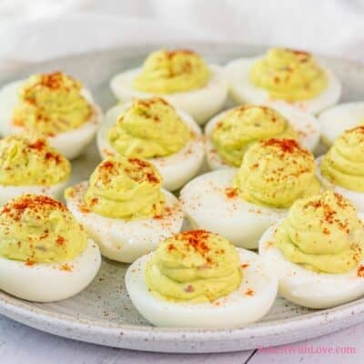 A close-up, slightly elevated shot shows a platter of avocado deviled eggs. Approximately a dozen halved hard-boiled egg whites are filled with a pale green, creamy avocado mixture, piped into a smooth, slightly mounded swirl. Each filled egg is lightly dusted with a sprinkle of reddish-orange paprika. The eggs are arranged on a light-colored, rustic-looking ceramic plate, against a soft, light-colored background.