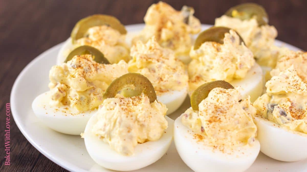 Plated jalapeno popper deviled eggs on white plate with halved slices of pickled jalapenos and a sprinkle of cayenne pepper on top.