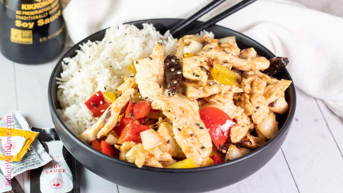A wide horizontal image showing tender wok-fried strips of chicken breast with peppers (firecracker chicken) ia black bowl over white rice with a set of chopsticks.