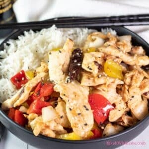 A square image showing tender wok-fried strips of chicken breast with peppers (firecracker chicken) in a black bowl over white rice with a set of chopsticks.