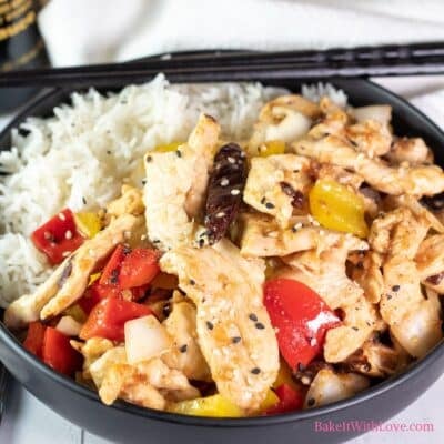 A square image showing tender wok-fried strips of chicken breast with peppers (firecracker chicken) in a black bowl over white rice with a set of chopsticks.