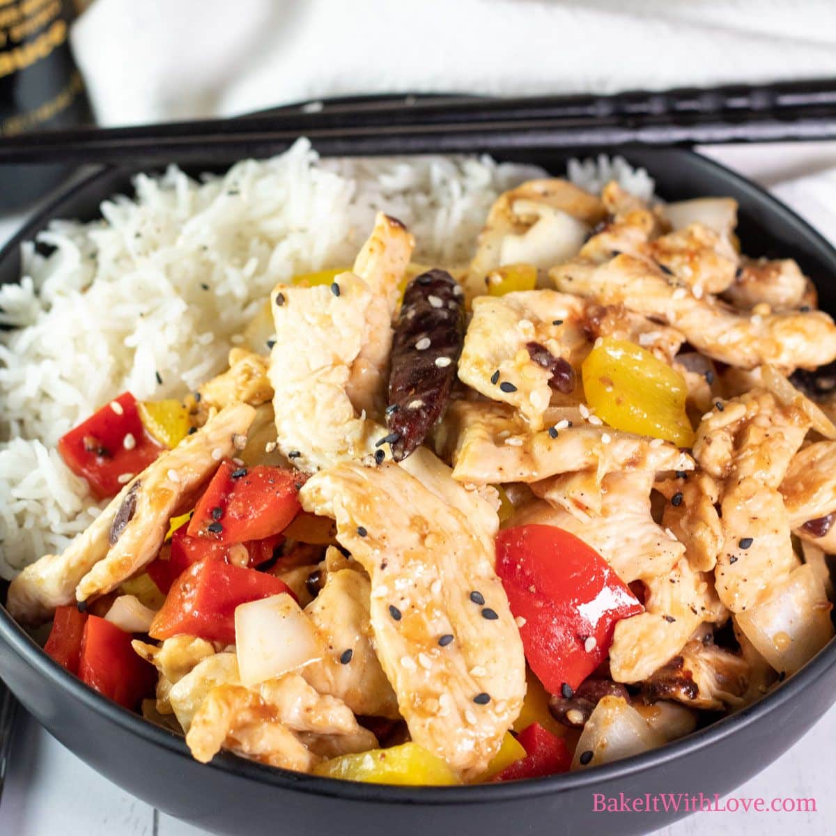 A square image showing tender wok-fried strips of chicken breast with peppers (firecracker chicken) in a black bowl over white rice with a set of chopsticks.