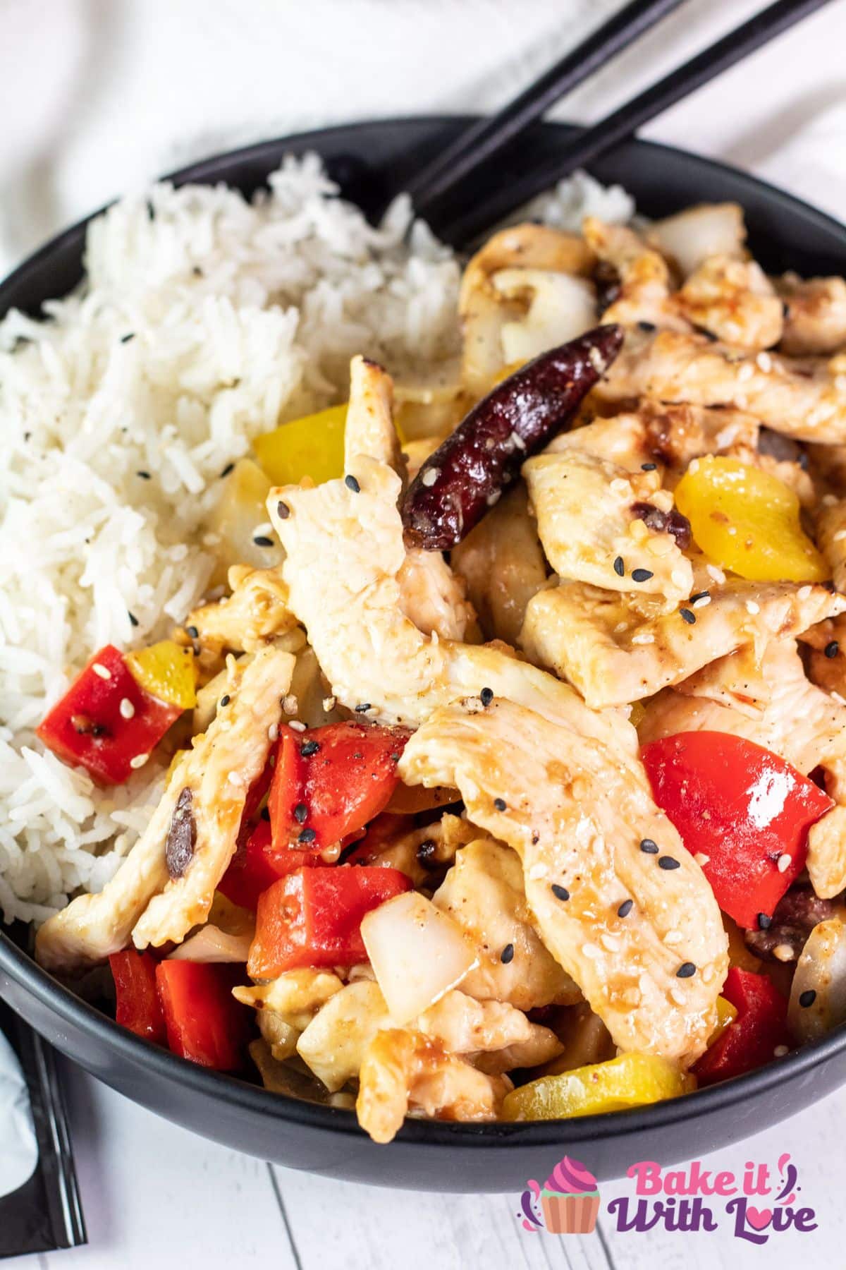 A tall image showing tender wok-fried strips of chicken breast with peppers (firecracker chicken) in a black bowl over white rice with a set of chopsticks.