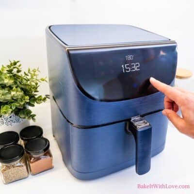 A close-up of a blue air fryer with a hand touching the digital display, which shows "180°" and "15:32". To the left, a green plant in a white pot and three small jars of spices are visible on a white counter. "BakeItWithLove.com" is at the bottom right.