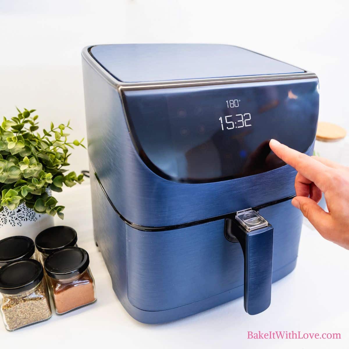 A close-up of a blue air fryer with a hand touching the digital display, which shows "180°" and "15:32". To the left, a green plant in a white pot and three small jars of spices are visible on a white counter. "BakeItWithLove.com" is at the bottom right.