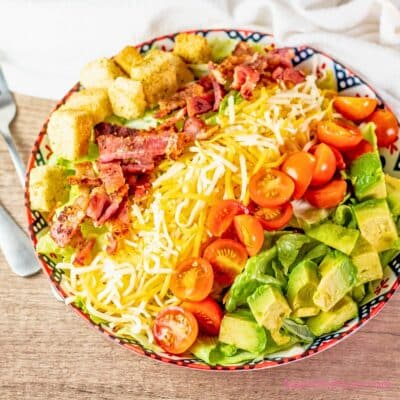 A square image of a clorful BLT salad with croutons, bacon, cheese, cherry tomatoes, and avocado on a wooden surface.