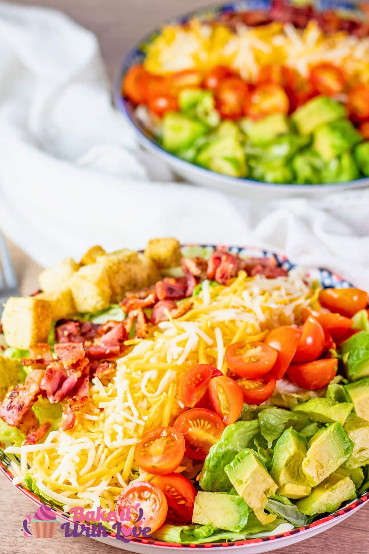 A tall image showing a colorful BLT salad with sections of cherry tomatoes, shredded cheese, bacon, croutons, and avocado in a bowl with the 'Bake It with Love' logo in the bottom left corner.